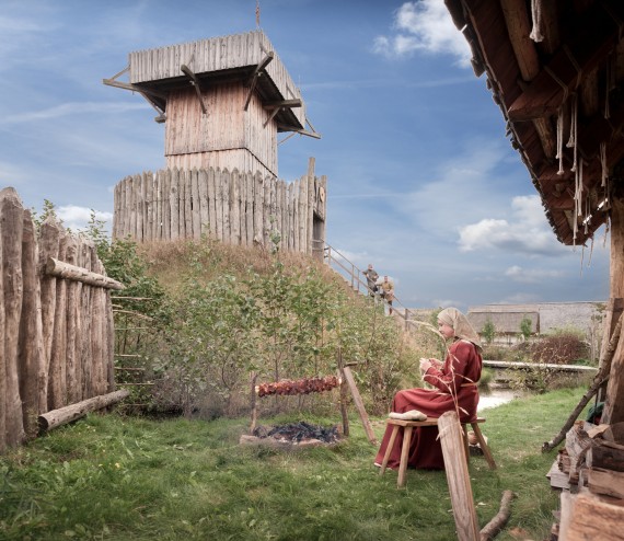 Im Vordergrund eine Frau in Mittelalterlicher Kleidung an einem Lagerfeuer. Im Hintergrund eine Turmhügelburg aus Holz