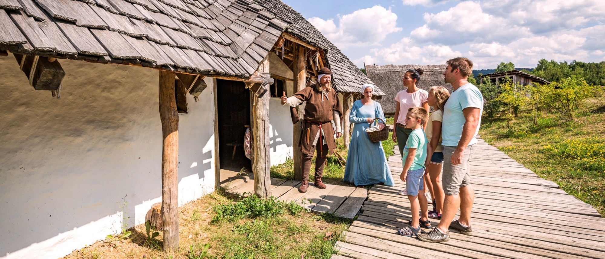 Zwei Personen in mittelalterlicher Kleidung zeigen einer Familie ein slawisches Haus aus Lehm und Holz